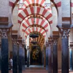 Perspectiva del interior de la Mezquita-Catedral de Córdoba, mostrando una nave de arcos de herradura bicolores (rojo y blanco) sostenidos por columnas de mármol y jaspe de tonos oscuros. Se aprecian las lámparas colgantes que iluminan el pasillo central, resaltando la arquitectura califal.