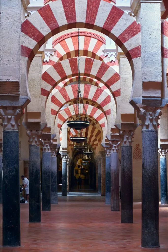 Perspectiva del interior de la Mezquita-Catedral de Córdoba, mostrando una nave de arcos de herradura bicolores (rojo y blanco) sostenidos por columnas de mármol y jaspe de tonos oscuros. Se aprecian las lámparas colgantes que iluminan el pasillo central, resaltando la arquitectura califal.