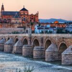 Vista panorámica del Puente Romano de Córdoba sobre el río Guadalquivir al atardecer, con la Mezquita-Catedral en el fondo y la Puerta del Puente a la izquierda. Se aprecian los arcos de piedra del puente y el reflejo de las luces naranjas y azules del cielo en el agua