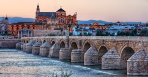 Vista panorámica del Puente Romano de Córdoba sobre el río Guadalquivir al atardecer, con la Mezquita-Catedral en el fondo y la Puerta del Puente a la izquierda. Se aprecian los arcos de piedra del puente y el reflejo de las luces naranjas y azules del cielo en el agua