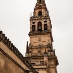 Primer plano en picado de la alta Torre Campanario de la Mezquita-Catedral de Córdoba, mostrando la verticalidad y la compleja estructura que combina el antiguo alminar islámico con la reedificación renacentista y barroca de piedra. Se aprecia la base de la muralla y parte de los tejados, sugiriendo su función como mirador.