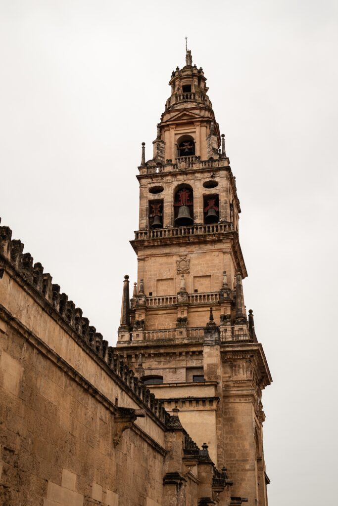 Primer plano en picado de la alta Torre Campanario de la Mezquita-Catedral de Córdoba, mostrando la verticalidad y la compleja estructura que combina el antiguo alminar islámico con la reedificación renacentista y barroca de piedra. Se aprecia la base de la muralla y parte de los tejados, sugiriendo su función como mirador.