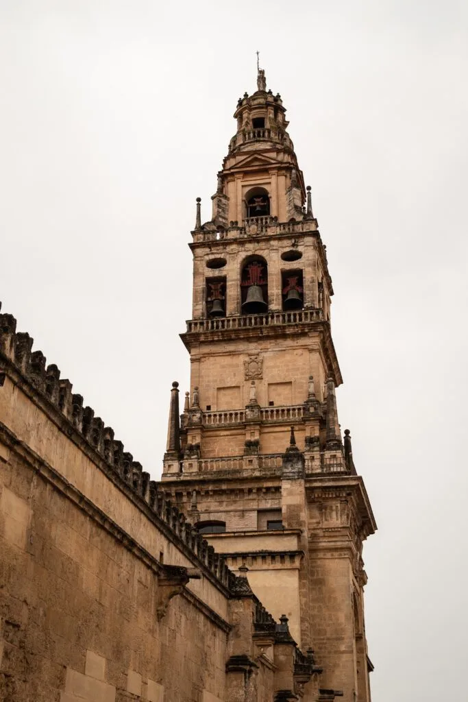 Primer plano en picado de la alta Torre Campanario de la Mezquita-Catedral de Córdoba, mostrando la verticalidad y la compleja estructura que combina el antiguo alminar islámico con la reedificación renacentista y barroca de piedra. Se aprecia la base de la muralla y parte de los tejados, sugiriendo su función como mirador.