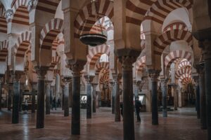 Vista interior del famoso Bosque de Columnas de la Mezquita-Catedral de Córdoba. La imagen muestra filas interminables de arcos de herradura bicolores (rojo y blanco) sostenidos por columnas de mármol y jaspe. Cuelgan grandes lámparas de bronce del techo.
