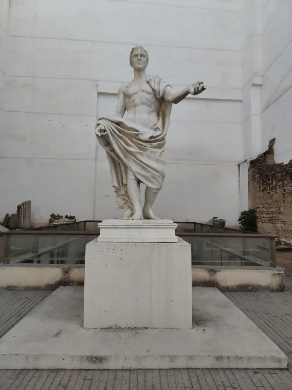 Fotografía de la estatua moderna de Marco Claudio Marcelo, un general romano, ubicada sobre un pedestal de piedra en la recepción del Templo Romano de Córdoba. La escultura, de mármol blanco, representa al general con una túnica drapeada alrededor de su cadera y con el brazo izquierdo extendido. Al fondo, una pared blanca lisa y restos de la antigua construcción romana.