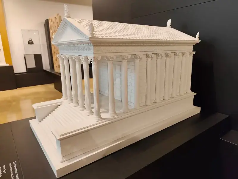Vista de una maqueta detallada y de color blanco del Templo Romano de Córdoba, exhibida en el Museo Arqueológico de la ciudad. La maqueta muestra la estructura completa del templo, incluyendo sus columnas corintias, el frontón triangular con relieves, el tejado a dos aguas y la escalinata de acceso. Se encuentra sobre una base negra de exposición y se aprecian otros objetos de museo al fondo.