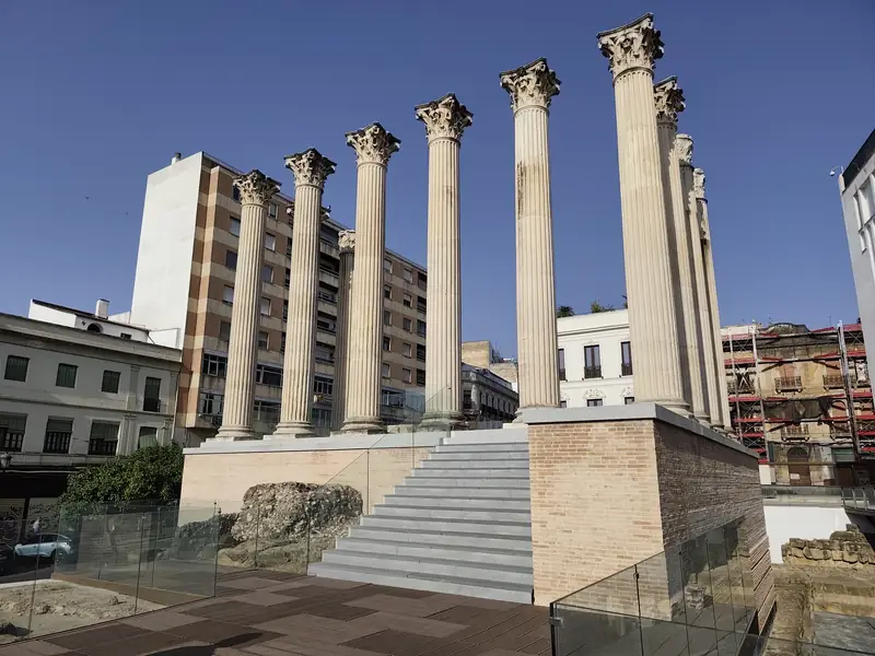 Vista frontal de las columnas del Templo Romano de Córdoba bajo, recién inaugurado para las visitas en junio de 2025. Se aprecian las escalinatas de acceso recién habilitadas y una plataforma de observación con barandillas de cristal que permite a los visitantes contemplar el yacimiento arqueológico, incluyendo los cimientos y partes del foro, rodeado de edificios modernos de la ciudad.