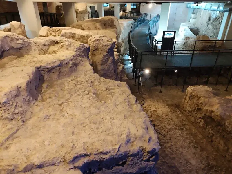 Vista de los restos arqueológicos del teatro romano de Córdoba, excavados bajo el Museo Arqueológico. La imagen muestra los cimientos de piedra y una pasarela metálica y acristalada que serpentea a través del yacimiento, permitiendo a los visitantes observarlo de cerca.
