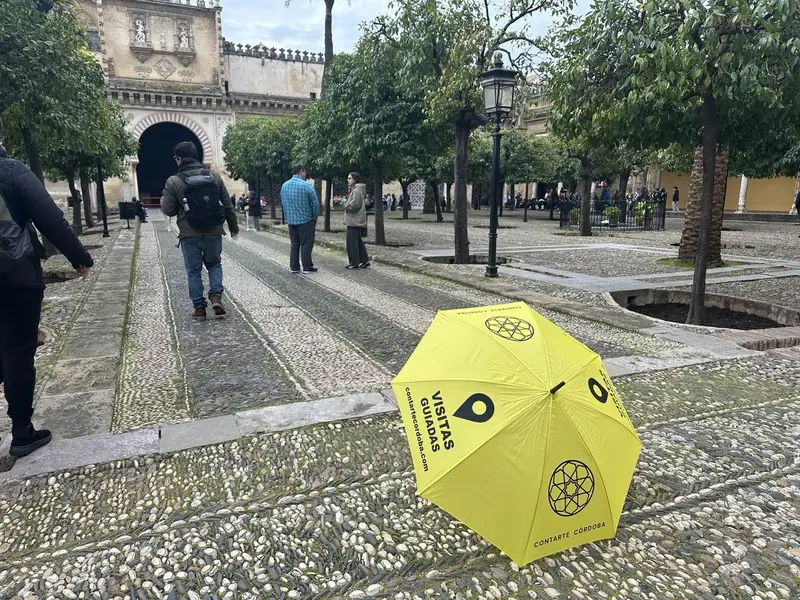 Vista a pie de calle del Patio de los Naranjos en la Mezquita de Córdoba, mostrando el empedrado tradicional, naranjos alineados y un paraguas amarillo de "Contarte Córdoba Visitas Guiadas" en primer plano junto a turistas y la Puerta de las Palmas al fondo.