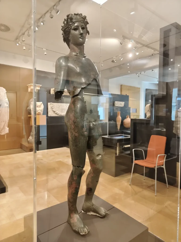 Estatua romana de bronce del Efebo Dionisíaco, con corona de hiedra y cuerpo fragmentado, exhibida en una vitrina del Museo Arqueológico de Córdoba. Se observan detalles de la técnica de fundición a la cera perdida.
