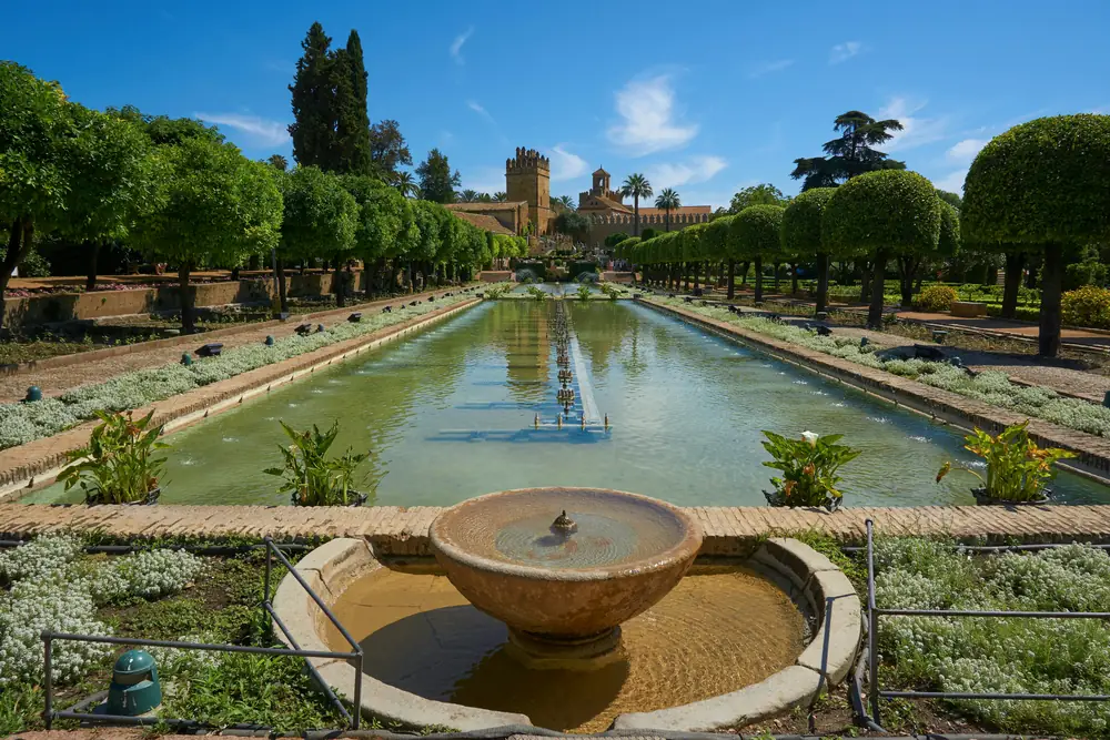 Vista panorámica de los estanques rectangulares del Alcázar de los Reyes Cristianos en Córdoba, flanqueados por hileras de naranjos podados en forma esférica y flores blancas, con la Torre de los Leones al fondo bajo un cielo azul despejado.