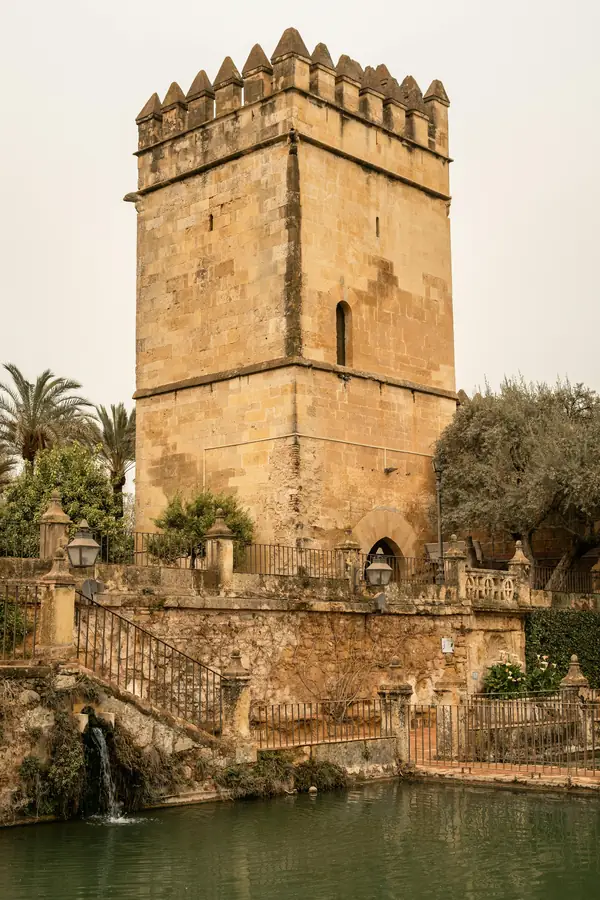 Vista lateral de la Torre de los Leones en el Alcázar de los Reyes Cristianos de Córdoba, destacando sus muros de sillería dorada, almenas escalonadas y una pequeña ventana de medio punto sobre un estanque de agua tranquila.