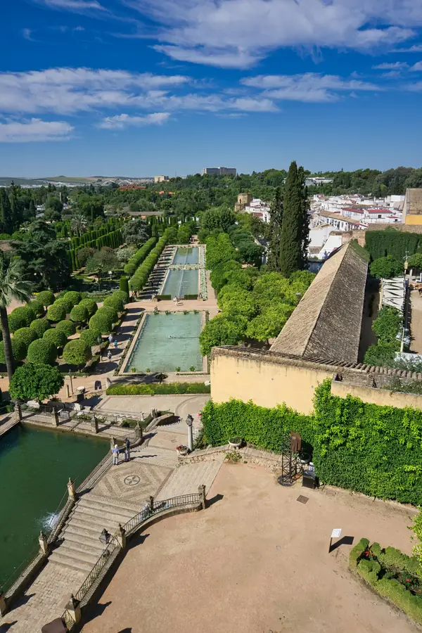 Fotografía desde una perspectiva elevada que muestra la extensión de los jardines del Alcázar de los Reyes Cristianos, con sus albercas rectangulares alineadas, setos de cipreses, naranjos en flor y las casas blancas de la Judería de Córdoba al fondo.