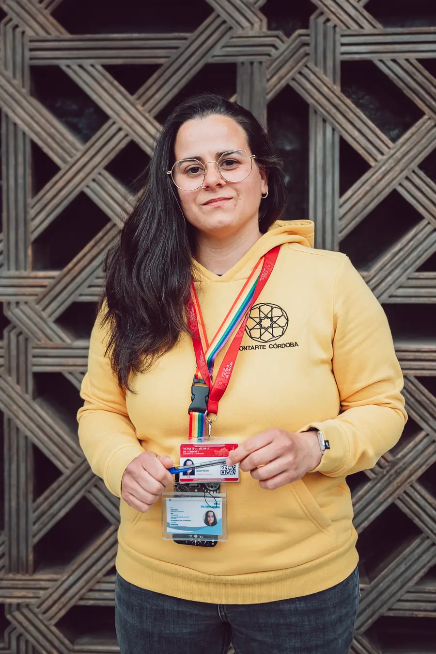 Adela, guía turístico oficial en Córdoba de Contarte Córdoba, posando con su acreditación oficial de intérprete de la Mezquita Catedral frente a la icónica celosía de madera diseñada por Rafael de la Hoz en la Mezquita-Catedral. La imagen destaca su carné de guía habilitada para ofrecer visitas guiadas profesionales.