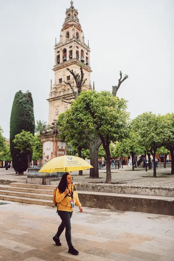 Adela Calzado, guía oficial de Contarte Córdoba, caminando por el Patio de los Naranjos con un paraguas amarillo corporativo. Al fondo destaca la Torre del Campanario de la Mezquita-Catedral de Córdoba bajo un cielo despejado.