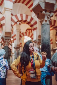 Adela Calzado, intérprete autorizada por la Mezquita Catedral de Córdoba, explicando la arquitectura e historia de la Mezquita. Al fondo se observa la perspectiva del bosque de columnas con los famosos arcos de herradura bicolores (rojo y blanco) y la iluminación tenue del monumento.